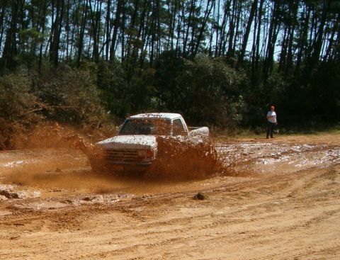 1989 Ford Bronco - A white pickup truck navigates a muddy track, kicking up a large spray of mud.