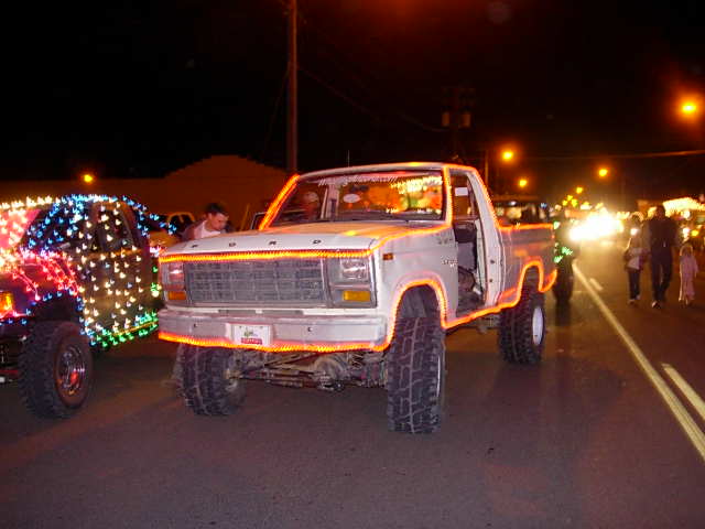 Front view of a white 1980-1986 Ford F-Series pickup truck decorated with orange and white lights, parked next to another truck covered in colorful lights