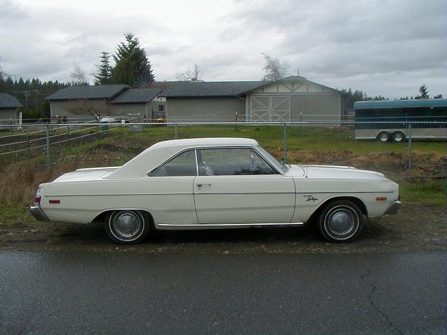 White coupe parked on asphalt, side view. Chrome trim, hubcaps, and a badge on the front fender are visible