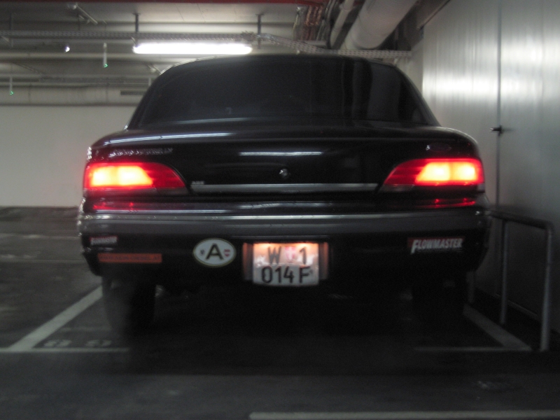 Rear of dark sedan in parking garage with Austrian plate, lit taillights, and Flowmaster bumper sticker