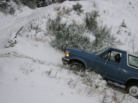 1992 Ford Bronco - A blue Ford Bronco navigates a snowy path, with snow-covered bushes and trees in the background.