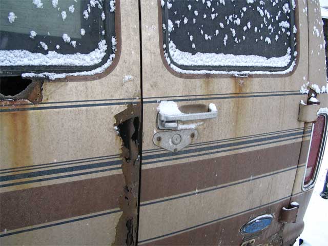 Rear doors of tan Ford van with heavy rust holes, snow on windows and handle