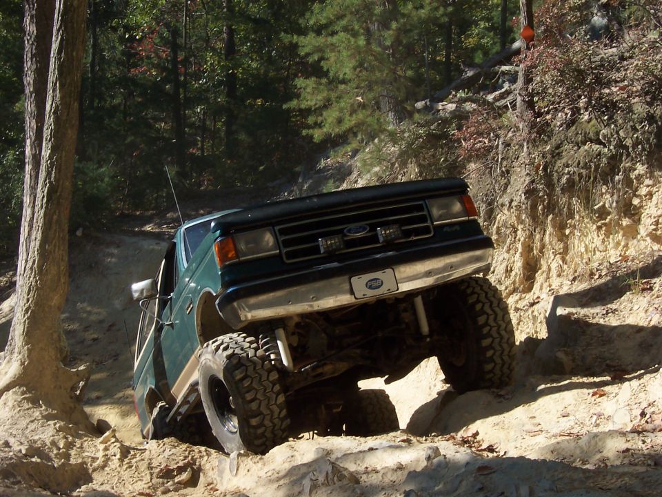 Green Ford pickup climbing a rocky off-road trail on oversized mud tires in the woods