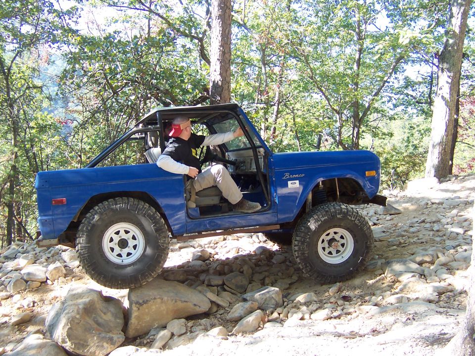 Blue early Ford Bronco with no top crawling rocks on a wooded trail, driver inside
