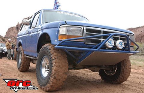 1984 Ford Bronco - A blue SUV, modified for off-road use, is parked on a dirt track with other vehicles in the background.
