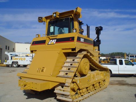 1966 International School Bus - A large yellow Caterpillar D9T bulldozer is parked outdoors, with other vehicles and buildings in the background under a blue sky.