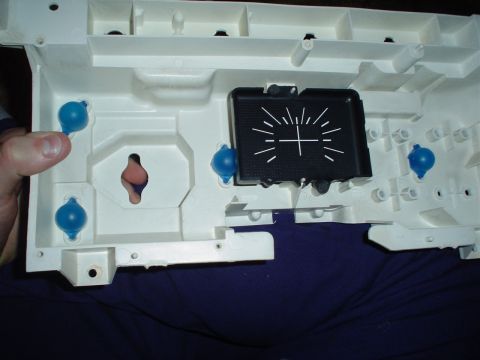 1984 Ford Bronco - Rear of a white plastic instrument cluster housing held on someone's lap, fitted with four blue-coated illumination bulbs in their sockets…