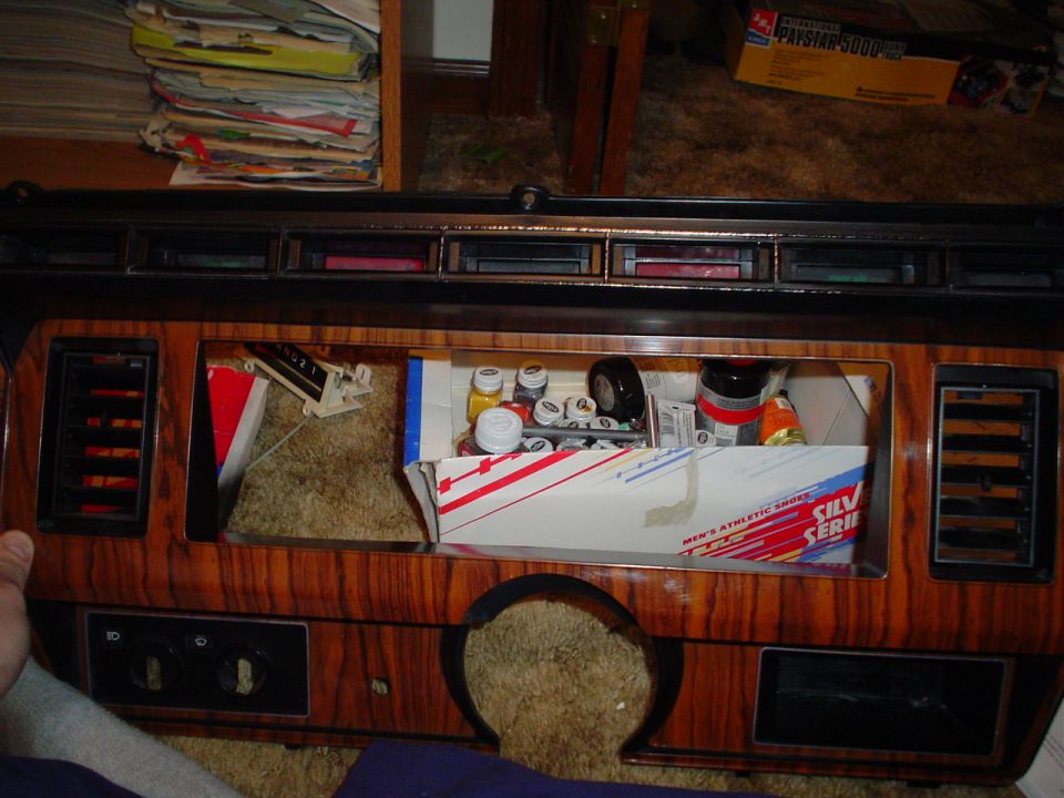 Dashboard with wood grain trim, open cubby filled with parts and a box of athletic shoes