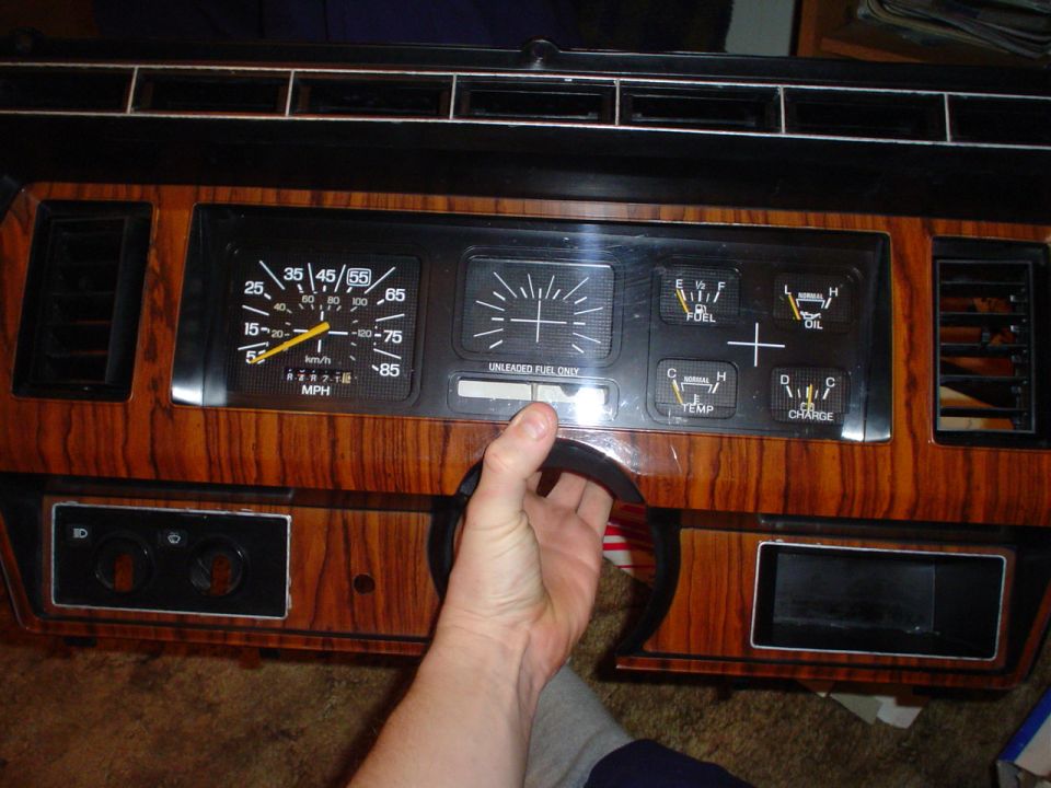 Dashboard with speedometer, fuel gauge, oil gauge, temperature gauge, and charge indicator. A hand is holding a part of the dashboard