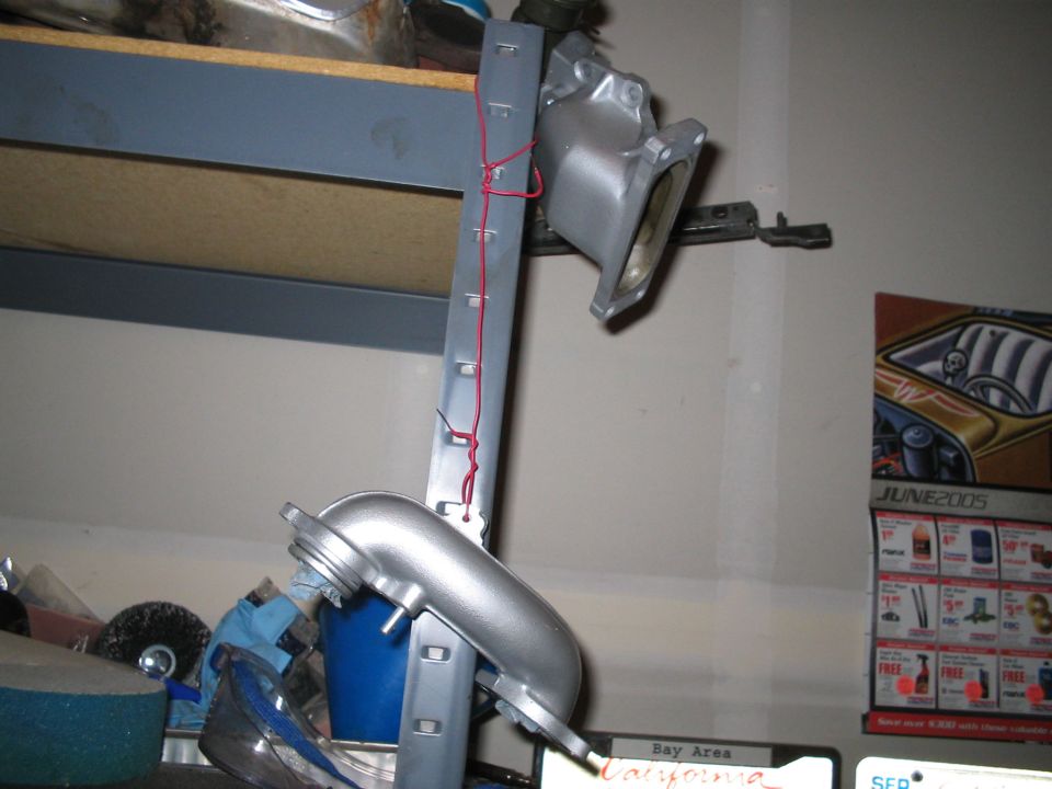 Silver metal engine intake manifold hanging from a gray metal strip with red wire