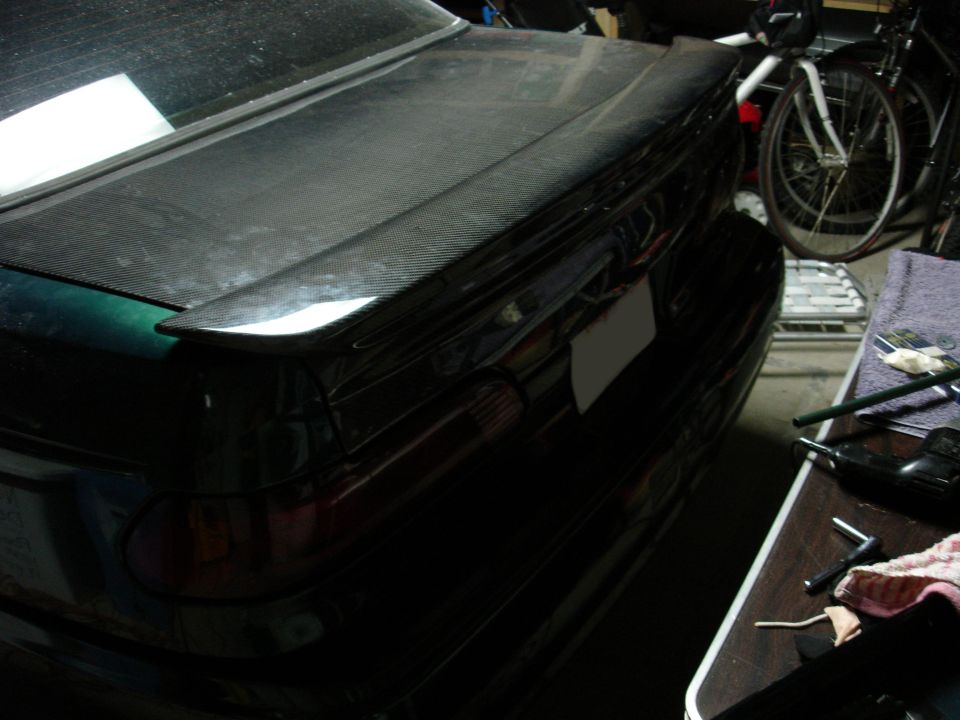 Rear of a dark sedan with a carbon fiber spoiler and a blank license plate