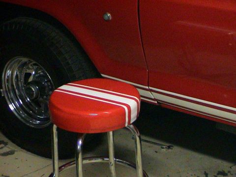 1979 Ford Bronco - A red vehicle with racing stripes is parked next to a stool.