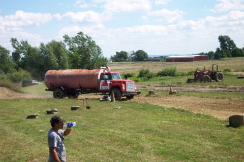 1979 Ford F-150 - A man drinks from a water bottle in front of a parked tanker truck and a tractor in a rural setting.