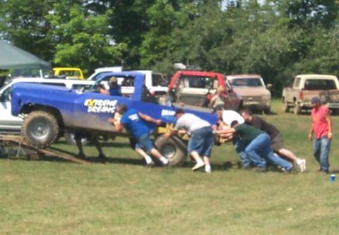 1979 Ford F-150 - Men push a blue pickup truck up a ramp during an event. Multiple other vehicles are present in the background.