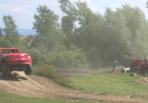 1979 Ford F-150 - A red truck is mid-air on a dirt track, with a tractor visible in the background.