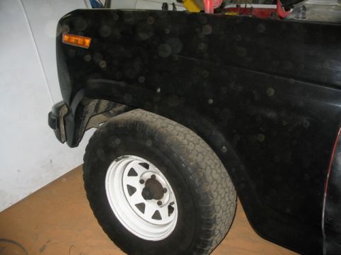 1981 Ford Bronco - The front corner of a black SUV is visible, showing the fender, headlight, and a tire with a white rim.