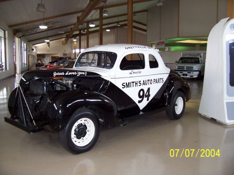 1987 Ford Bronco - A vintage coupe race car is displayed indoors with other vehicles in the background.