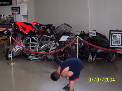 1987 Ford Bronco - A severely damaged race car is displayed behind a velvet rope, with a young boy observing it.