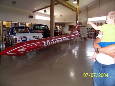 1987 Ford Bronco - A Budweiser dragster is displayed indoors next to a race truck, with a child pointing at it.