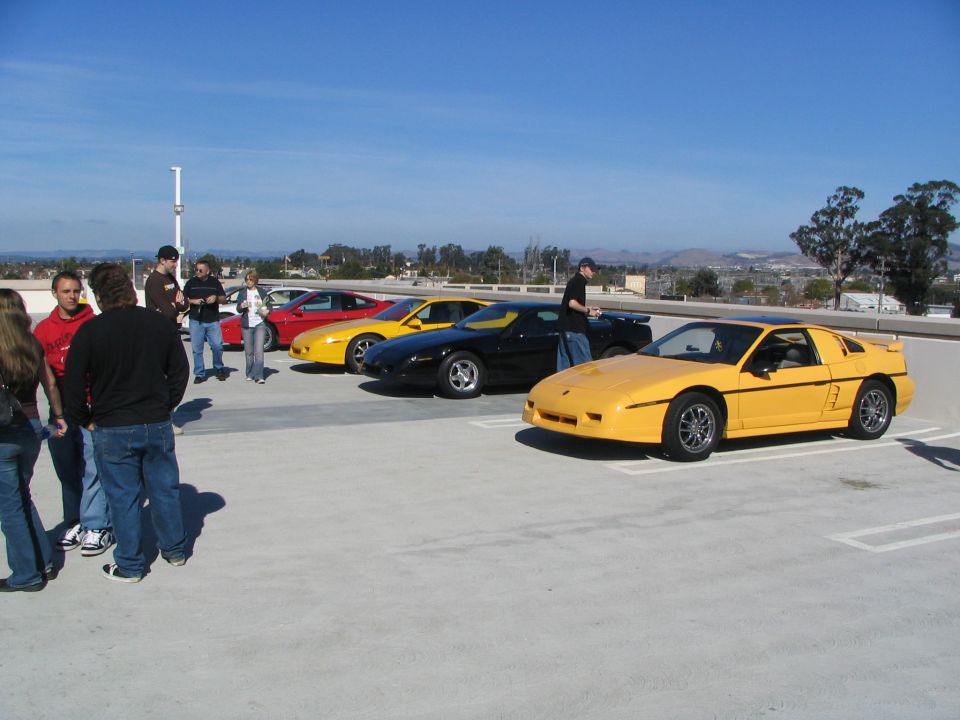 Yellow 1986 Pontiac Fiero parked in front of other Fieros and people