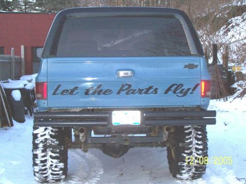 1979 Ford F-150 - A blue pickup truck with a camper shell and "Let the Parts Fly!" decal is seen from the rear in a snowy environment.
