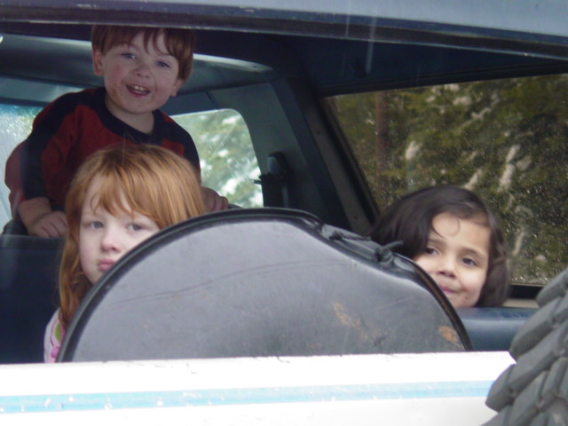 Three young children peering out the open side window of a vehicle, smiling at the camera