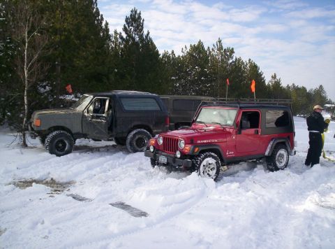 1996 Ford Bronco - Two off-road vehicles are parked in a snowy landscape, with a line of trees behind them.