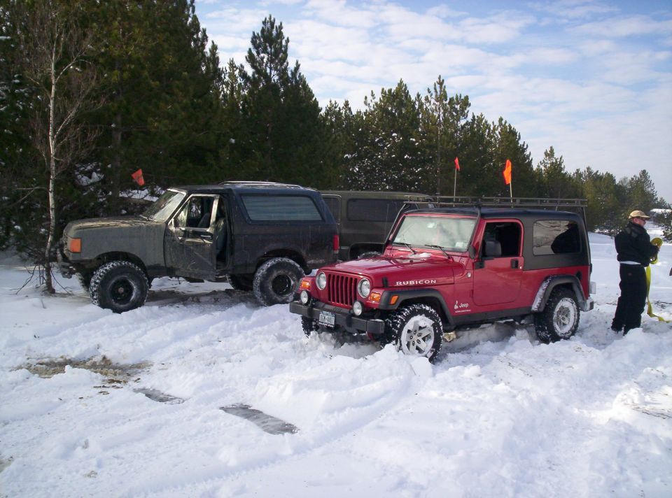 Red Jeep Wrangler and a dark SUV parked in the snow with trees in the background