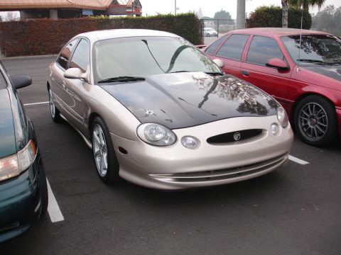 1995 Ford Taurus - A gold Ford Taurus with a black hood is parked in a lot next to other vehicles.
