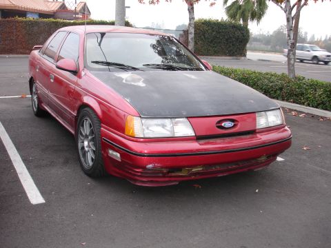1995 Ford Taurus - A red sedan with a matte black hood and damaged front bumper is parked in a lot.