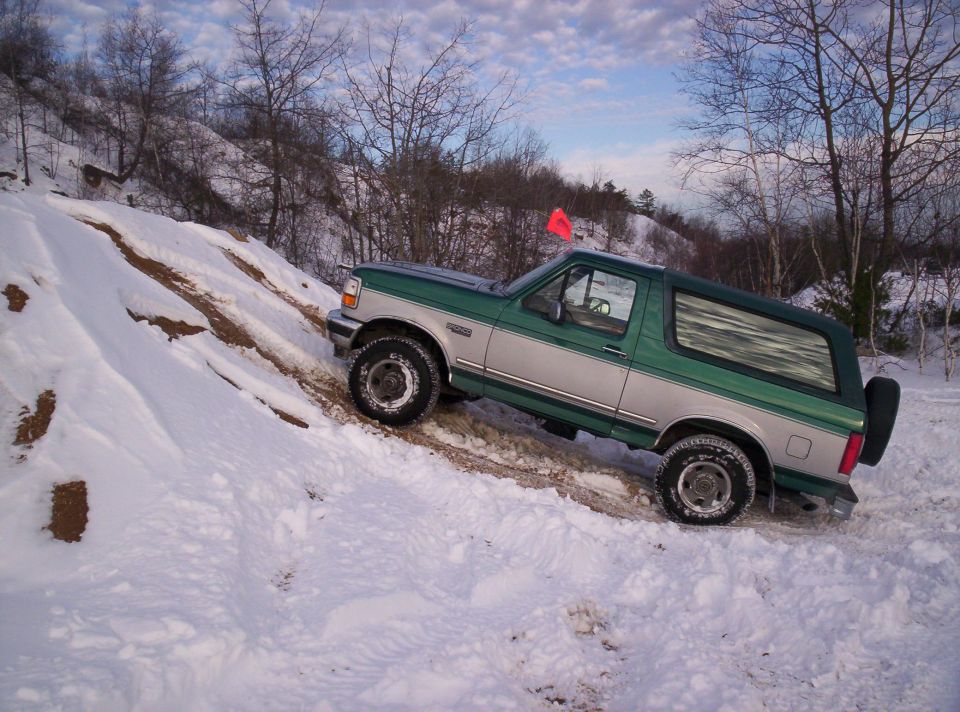 1996 Ford Bronco driving up a snowy, rutted hill with trees in the background