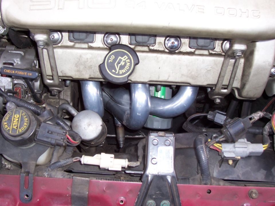 Engine bay close-up showing exhaust manifold, oil cap, and various hoses and connectors