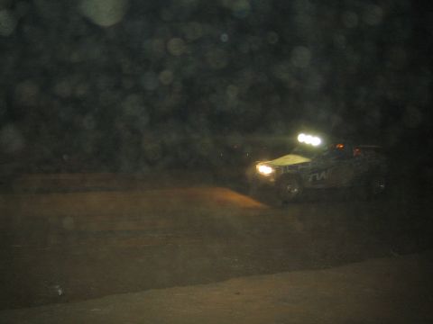 1990 Ford Bronco - distant off-road race truck with a glowing roof light bar drives across a dark dusty desert course at night.