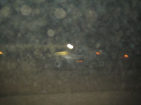 1990 Ford Bronco - dark off-road race car streaks in motion blur across a dusty desert course at night, its headlight glowing.