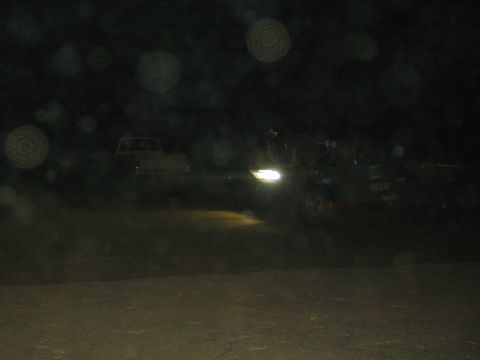 1990 Ford Bronco - off-road race vehicle with a bright headlight sits beside a parked car on a dark dusty desert course at night.