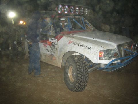 1990 Ford Bronco - white off-road race truck with a bank of roof lights and a Yokohama decal sits in a lit pit at night as crew work nearby.