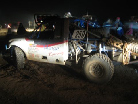 1990 Ford Bronco - white off-road race truck marked Mango Racing and number 250 with large mud tires sits in a churned-dirt pit at night.