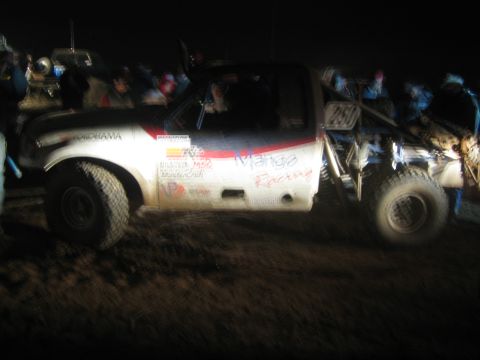 1990 Ford Bronco - mud-spattered white race truck marked 'Mango Racing' sits among a nighttime crowd at an off-road event.