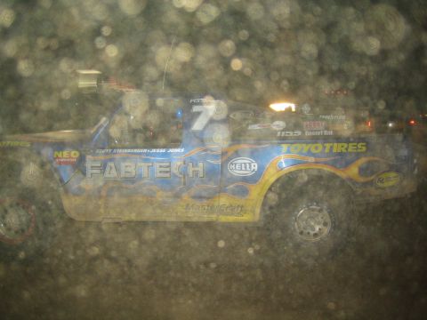 1990 Ford Bronco - blue number-7 trophy truck wearing Fabtech and Toyo Tires livery, viewed at night through a spotted lens.