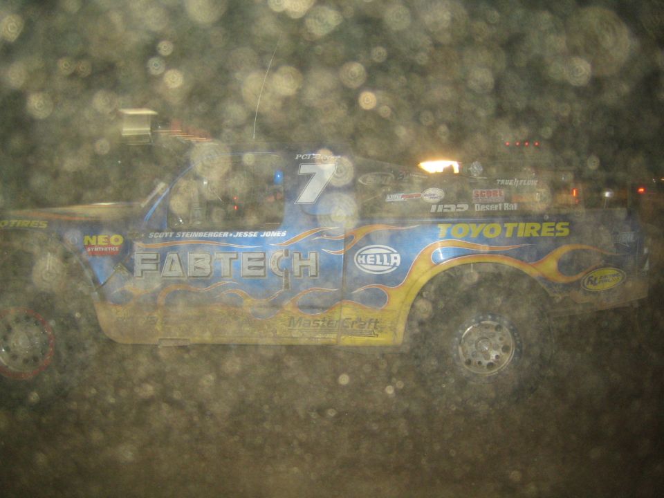 Blue trophy truck with 'Fabtech' and 'Toyo Tires' livery, number 7, shot through dirty glass