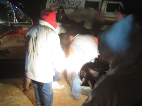 1990 Ford Bronco - Crew members in winter jackets gather and work in the dark beside a 'Mango Racing' off-road truck.