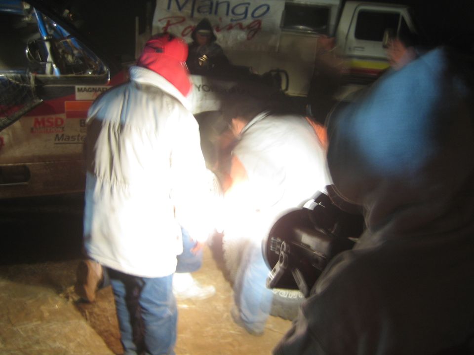 People in jackets crouched working at night beside a 'Mango Racing' race truck