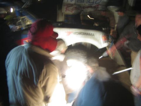 1990 Ford Bronco - motion-blurred night shot of people clustered around the wheel of a white Mango Racing off-road truck.