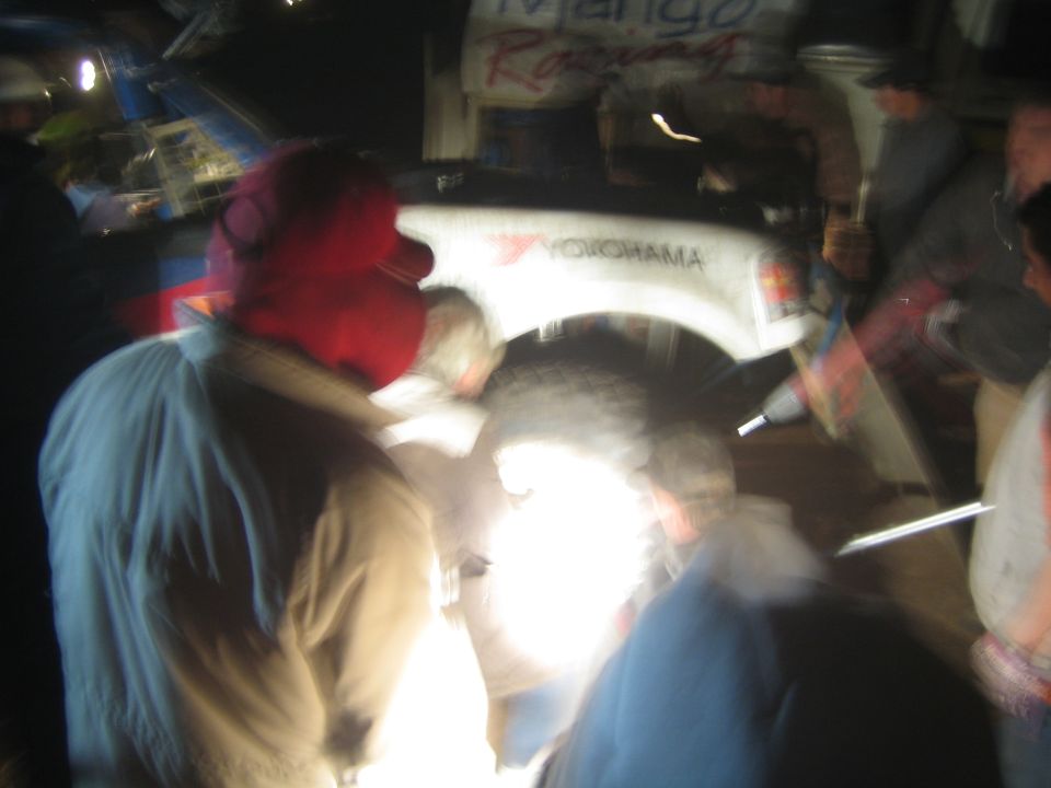 Blurry night scene of people crowded around a white 'Mango Racing' race truck wheel