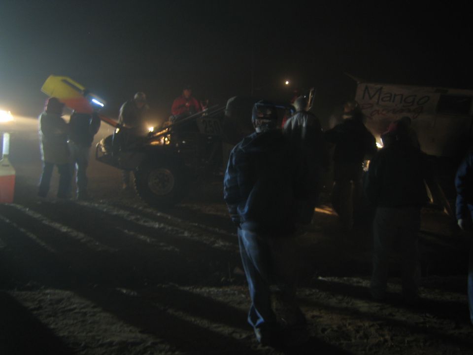 People standing in a foggy night desert pit area with a race vehicle and banner