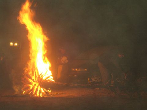 1990 Ford Bronco - tall bonfire blazes at night while people and a vehicle are silhouetted in the smoky darkness behind.