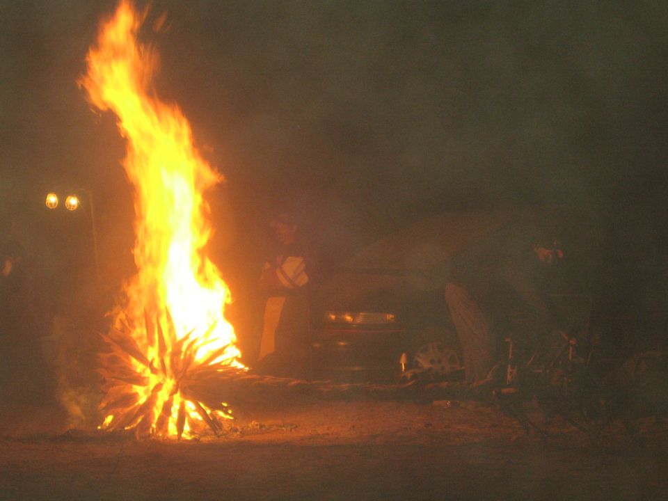Large open bonfire at night with people and a vehicle silhouetted behind