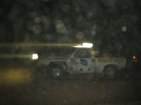 1990 Ford Bronco - white off-road race truck marked 'Best Buy' speeds through the night with its light bar illuminated.