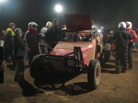 1990 Ford Bronco - Spectators in helmets gather around a red number-1207 off-road buggy by BFGoodrich Racing banners at night.