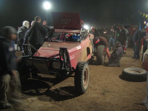 1990 Ford Bronco - pit crew works to change a tire on the red number-1207 off-road buggy under lights at night.